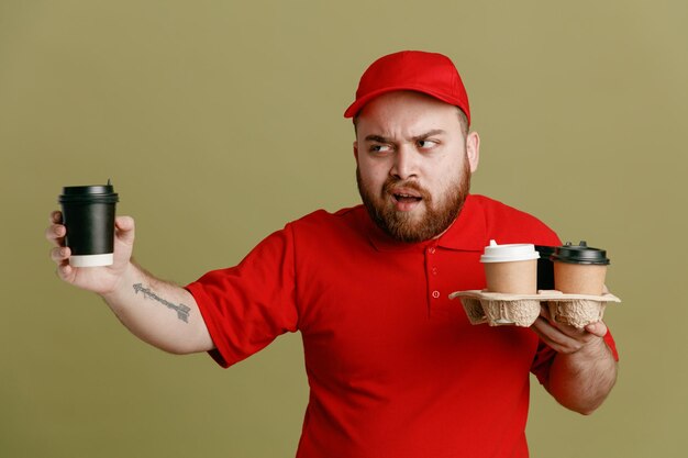 Empleado repartidor en uniforme de camiseta en blanco con gorra roja sosteniendo tazas de café mirando a un lado disgustado y enojado de pie sobre fondo verde