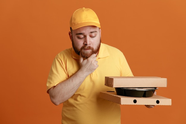 Empleado de reparto con uniforme de camiseta en blanco con gorra amarilla sosteniendo un contenedor de comida y cajas de pizza mirando cajas con expresión pensativa de pie sobre fondo naranja