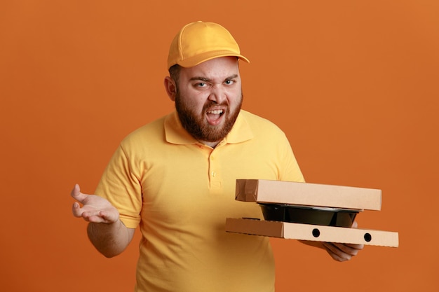 Empleado de reparto con uniforme de camiseta en blanco con gorra amarilla sosteniendo un contenedor de comida y cajas de pizza mirando a la cámara con cara enojada de pie sobre fondo naranja