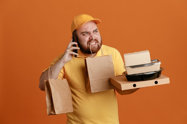 Empleado de reparto con uniforme de camiseta en blanco con gorra amarilla sosteniendo un recipiente de comida y una caja de pizza con una bolsa de papel en los dientes hablando por teléfono móvil con expresión molesta de pie sobre un fondo naranja