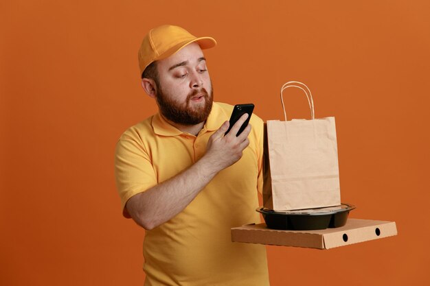 Empleado de reparto con uniforme de camiseta en blanco con gorra amarilla sosteniendo un recipiente de comida y una caja de pizza con una bolsa de papel mirando el teléfono inteligente con cara seria de pie sobre fondo naranja