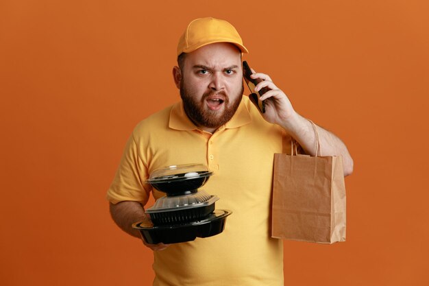 Empleado de reparto con uniforme de camiseta en blanco con gorra amarilla sosteniendo recipientes de comida con una bolsa de papel hablando por teléfono móvil molesto e irritado de pie sobre fondo naranja