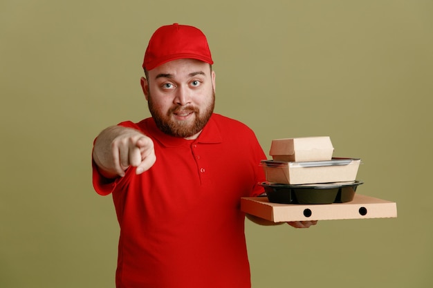 Empleado de reparto con uniforme de camiseta en blanco con gorra roja sosteniendo contenedores de comida y caja de pizza apuntando con el dedo índice a la cámara sonriendo amigablemente de pie sobre fondo verde