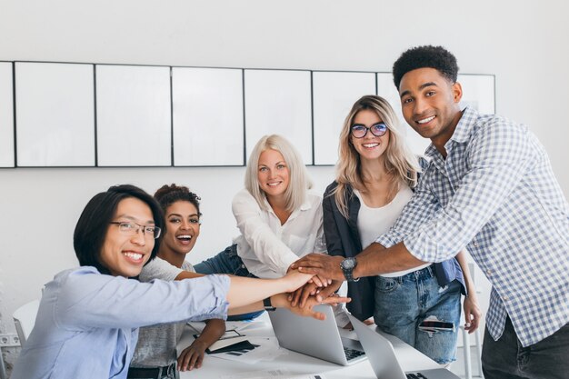 Empleado rubio encantador que se divierte con sus colegas y que posa para la foto en la sala de luz. El equipo de especialistas en TI puso fin a un gran proyecto empresarial y un apretón de manos.