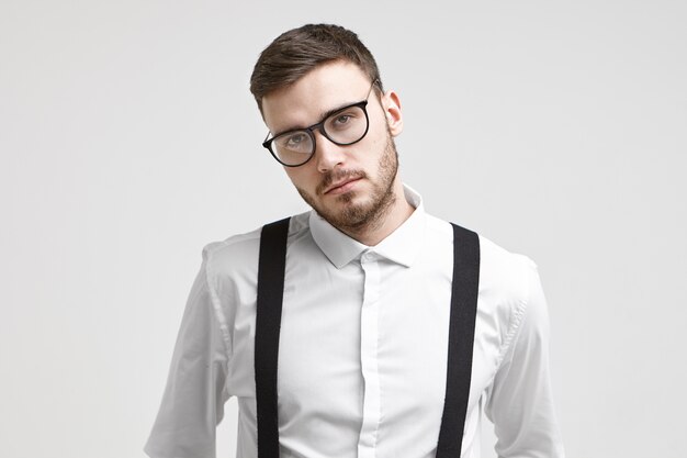 Empleado de sexo masculino sin afeitar joven hermoso serio que lleva anteojos de moda y camisa formal blanca con tirantes que presenta aislada contra el fondo blanco de la pared del estudio con copyspace para su información