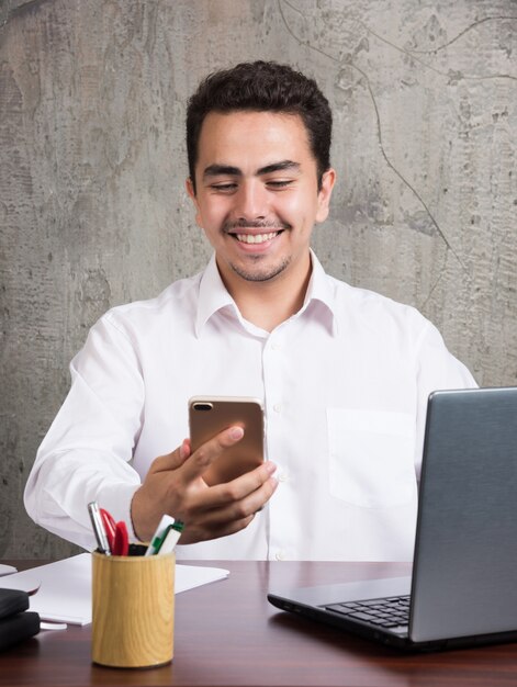 Empleado sonriente mirando por teléfono y sentado en el escritorio. Foto de alta calidad