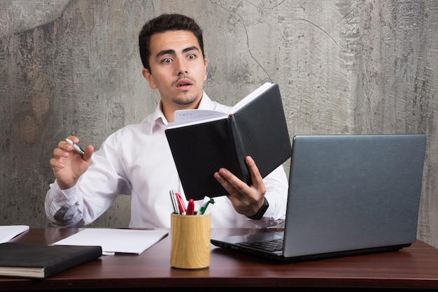 Empleado sorprendido mirando en el cuaderno y sentado en el escritorio. Foto de alta calidad