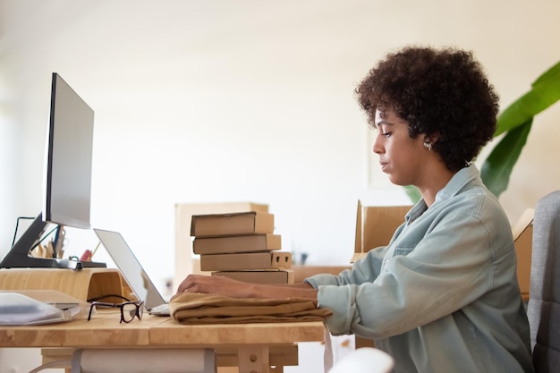 Foto gratuita empleado de tienda en línea usando laptop. una mujer de negocios afroamericana seria sentada a la mesa con un montón de paquetes. concepto de logística