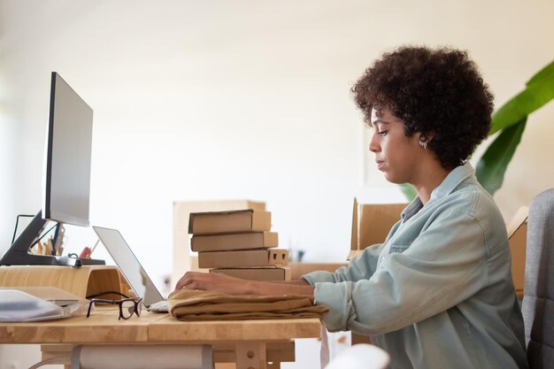 Empleado de tienda en línea usando laptop. Una mujer de negocios afroamericana seria sentada a la mesa con un montón de paquetes. Concepto de logística