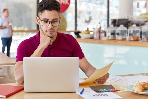 El empleador masculino creativo hace trabajo remoto en el interior de la cafetería, se viste informalmente, descarga archivos de Internet en una computadora portátil, está rodeado de papeles, tiene una carrera exitosa, come en la cafetería