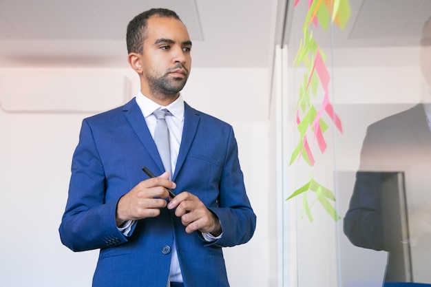 Empleador de oficina afroamericano sosteniendo la pluma y leyendo notas en la pared de vidrio. Hombre de negocios confiado enfocado en traje pensando en la idea para el proyecto