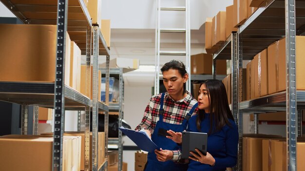 Empleados asiáticos revisando los suministros del almacén en tabletas y archivos, trabajando con la logística de la sala de almacenamiento. Equipo joven de personas que usan artilugios y papeles para el plan de negocios y la producción.