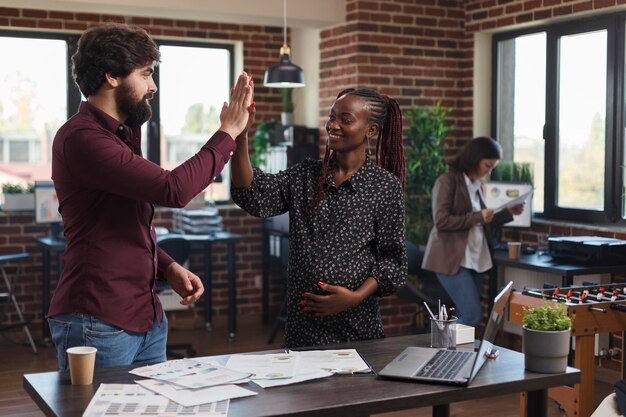 Empleados de la empresa de marketing felicitándose unos a otros por los excelentes resultados de la estrategia de marketing de inicio. Feliz sonriente mujer embarazada choca los cinco con su compañero de trabajo para ideas de presentación increíbles.