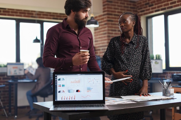 Empleados de empresas multiétnicas revisando los ingresos anuales de la empresa y discutiendo la estrategia de marketing de inicio. Compañeros de trabajo de la agencia financiera intercambian ideas de proyectos con una computadora portátil que muestra gráficos en pantalla.