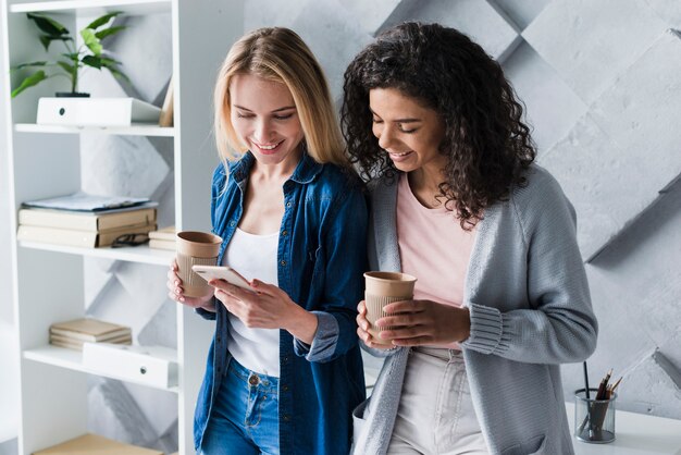 Empleados de oficina étnicos femeninos en el descanso para tomar café con teléfono inteligente