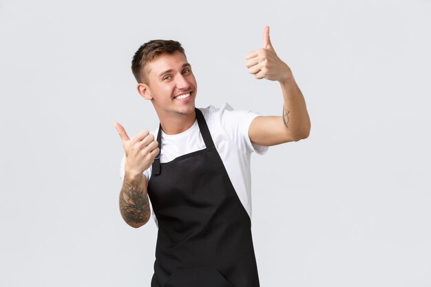 Empleados, tiendas de abarrotes y concepto de cafetería. Barista amable guapo en delantal negro, mostrando el pulgar hacia arriba, invitando a capuchino, sonriendo encantado, garantía de calidad, fondo blanco.