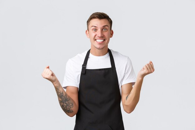 Empleados, tiendas de abarrotes y concepto de cafetería. Sonriendo barista guapo satisfecho en delantal negro celebrando el éxito. Bomba de puño de camarero alegre y gritando sí en regocijo, fondo blanco.