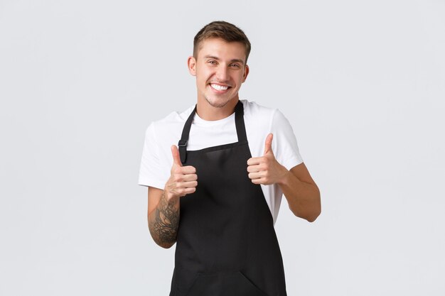 Empleados, tiendas de abarrotes y concepto de cafetería. Trabajador guapo y amable en la cafetería, empleado de la tienda con delantal negro, mostrando el pulgar hacia arriba y sonriendo para dar la bienvenida a los huéspedes, garantía de calidad