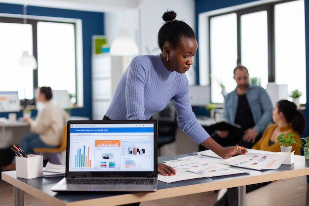 Foto gratuita emprendedor africano de la puesta en marcha de la empresa leyendo gráficos en el papeleo de los documentos. equipo diverso de gente de negocios que analiza los informes financieros de la empresa desde la computadora. ent profesional empresarial exitoso