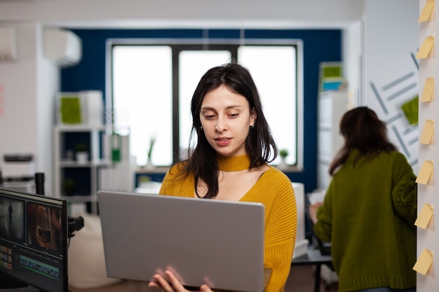 Emprendedor enfocado de pie en la oficina de la agencia creativa sosteniendo una computadora portátil y escribiendo información del proyecto