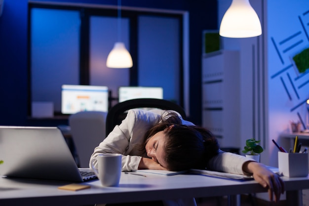 Emprendedor que trabaja horas extras en un proyecto de marketing, quedándose dormido en el escritorio mientras mira documentos financieros tratando de respetar la fecha límite