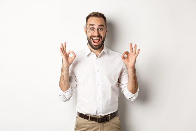 Emprendedor sorprendido que muestra el signo correcto y parece feliz, satisfecho con el producto, de pie blanco