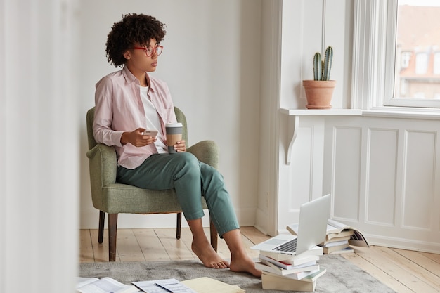 Una emprendedora femenina exitosa y reflexiva disfruta de una bebida de capuchino, sostiene una taza para llevar, se sienta en un sillón, usa un teléfono celular y una computadora portátil