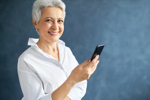 Foto gratuita emprendedora de mediana edad con cabello gris corto sosteniendo móvil, haciendo llamadas de negocios, escribiendo mensajes de texto.