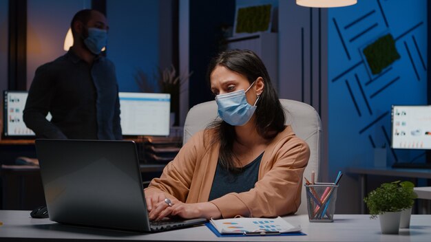 Empresaria adicta al trabajo con máscara facial contra covid19 trabajando en la oficina de inicio analizando la estrategia económica a altas horas de la noche