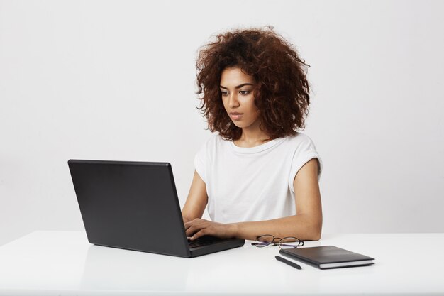 Empresaria africana hermosa que mira la computadora portátil sobre la pared blanca.