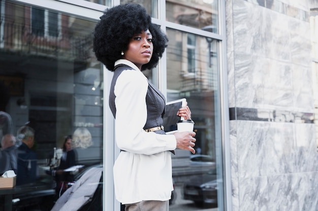 Empresaria africana joven confiada que se coloca fuera de la oficina que sostiene la tableta digital y la taza de café disponible