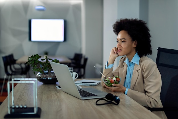 Empresaria afroamericana navegando por la red en una computadora mientras come ensalada en la oficina