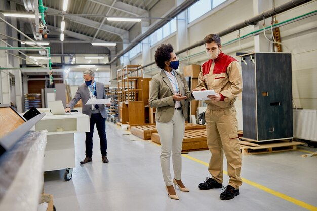 Empresaria afroamericana y trabajador masculino con máscaras faciales mientras examina informes en una fábrica de madera