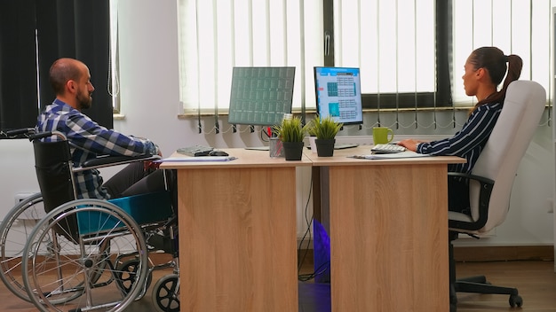 Foto gratuita empresaria analizando estadísticas financieras hablando con un compañero de trabajo discapacitado sentado en silla de ruedas, control de gráficos en el escritorio en la oficina del edificio. empresario discapacitado con tecnología moderna