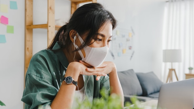 Empresaria de Asia con mascarilla médica usando laptop hablar con colegas sobre el plan en videollamada mientras trabaja desde casa en la sala de estar.