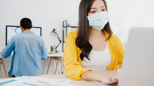 Empresaria de Asia que lleva una mascarilla médica para el distanciamiento social en una nueva situación normal para la prevención de virus mientras usa la computadora portátil en el trabajo en la oficina. Estilo de vida después del virus corona.