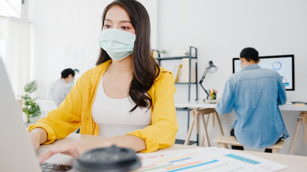 Empresaria de Asia que lleva una mascarilla médica para el distanciamiento social en una nueva situación normal para la prevención de virus mientras usa la computadora portátil en el trabajo en la oficina. Estilo de vida después del virus corona.