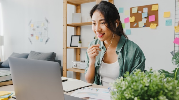 La empresaria de Asia que usa la computadora portátil habla con sus colegas sobre el plan en la videollamada mientras trabaja de manera inteligente desde casa en la sala de estar.