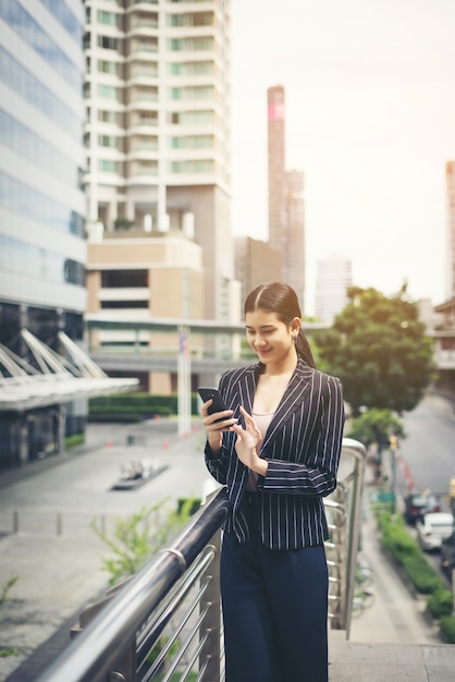 Empresaria asiática joven que usa en smartphone.Young móvil profesional en la ciudad delante de edificio grande.