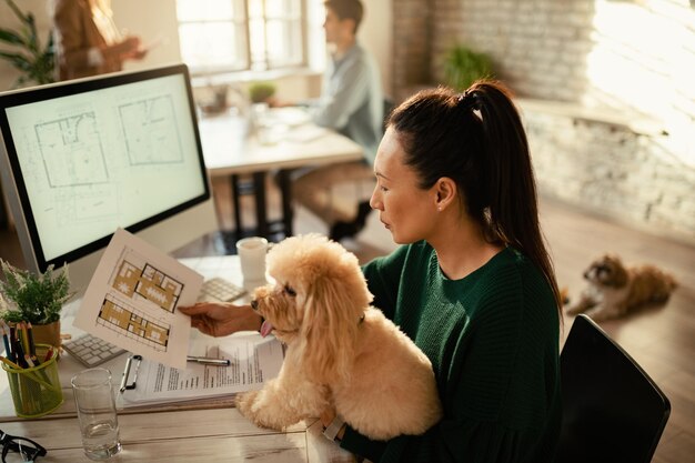 Empresaria asiática leyendo planos mientras sostiene a su perro y trabaja en la oficina