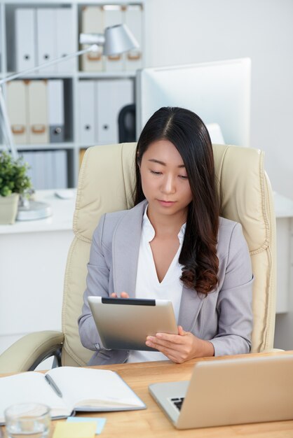 Empresaria asiática que se sienta en el escritorio en oficina y que usa la tableta
