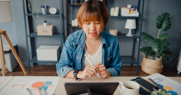 La empresaria asiática que usa la tableta hable con sus colegas sobre el plan en una videollamada mientras trabaja desde su casa en la sala