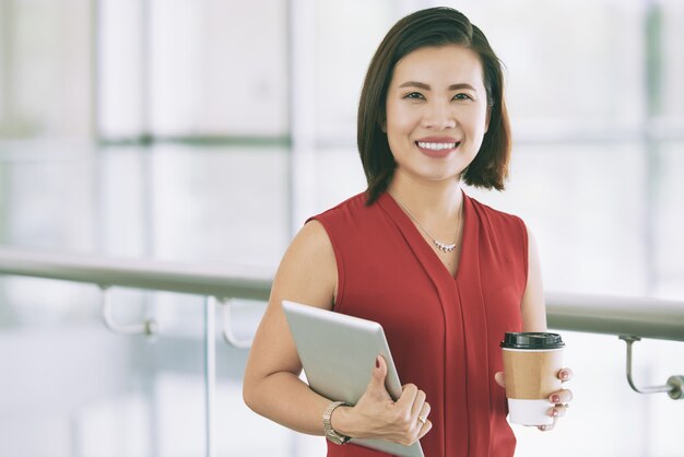 Empresaria asiática sonriente posando en el interior en el balcón con tableta y café para llevar