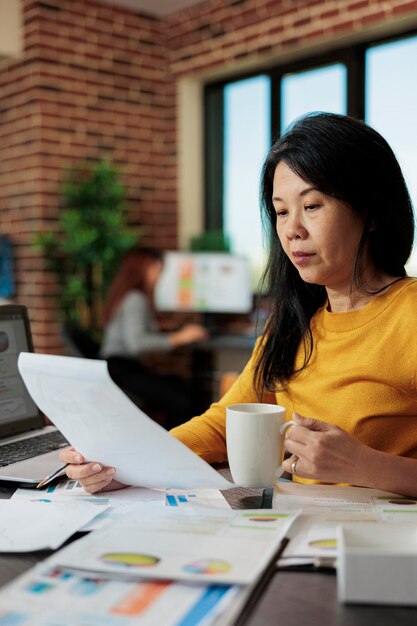 Empresaria asiática sosteniendo un portapapeles con estadísticas de marketing trabajando en la estrategia empresarial en la oficina de inicio. Gerente ejecutivo analizando gráficos, lluvia de ideas para el aumento de la empresa