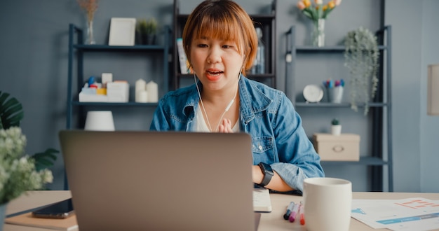 La empresaria asiática usando una computadora portátil hable con sus colegas sobre el plan en una videollamada mientras trabaja desde su casa en la sala de estar