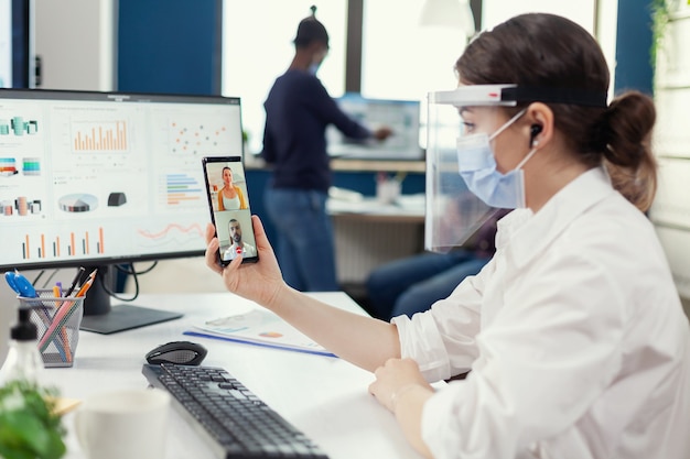 La empresaria con auriculares inalámbricos durante la conferencia en línea en el teléfono inteligente con mascarilla en el lugar de trabajo. Compañeros de trabajo multiétnicos que trabajan respetando la distancia social en los negocios durante la pandemia global