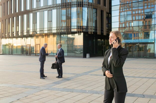 Empresaria concentrada vistiendo traje de oficina, hablando por teléfono móvil al aire libre. Los empresarios y la fachada de cristal del edificio de la ciudad en segundo plano. Copie el espacio. Concepto de comunicación empresarial