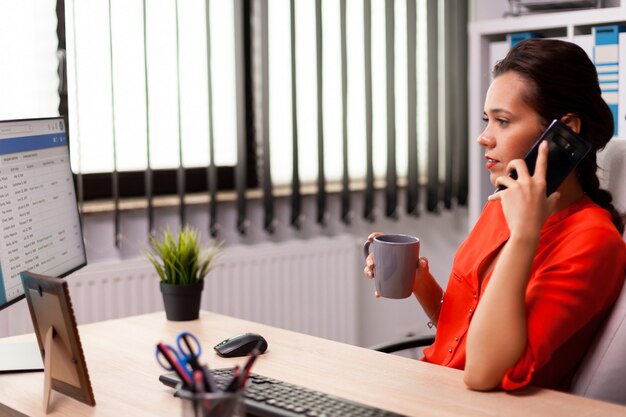 Empresaria corporativa en el lugar de trabajo hablando por teléfono con un socio de negocios vistiendo de rojo. Freelancer ocupado que trabaja con un teléfono inteligente desde la oficina para hablar con los clientes sentados en el escritorio mirando el documento.