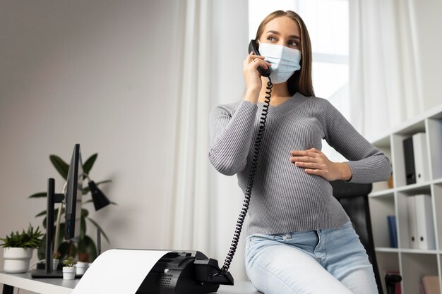 Empresaria embarazada con máscara médica tomando llamadas en la oficina