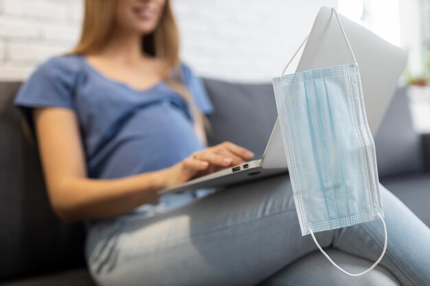 Empresaria embarazada en el sofá trabajando en equipo portátil con máscara médica